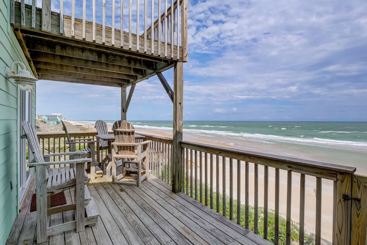 A tranquil oceanfront deck features classic adirondack chairs facing out toward the water, with the sandy beach and gentle waves clearly visible. The space feels inviting for both humans and pets to relax, enjoy ocean breezes, and watch the sunrise or sunset together.